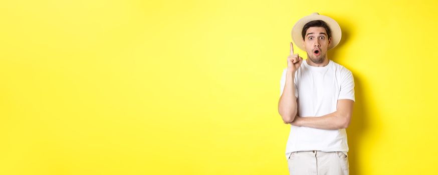 Portrait of young man in straw hat having an idea, raising finger eureka sign, making suggestion, standing over yellow background.