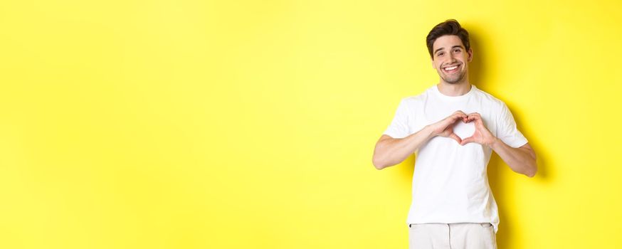 Happy romantic man showing heart sign, smiling and express love, standing over yellow background. Copy space