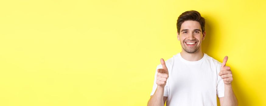 Confident man pointing fingers at camera and smiling, praising you, standing over yellow background.