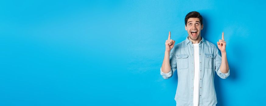 Excited handsome man pointing and looking up, showing advertisement, standing over blue background.