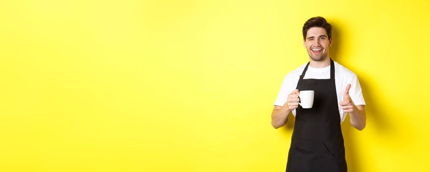 Handsome barista in black apron holding coffee cup, pointing finger at you, inviting visit cafe, standing over yellow background.