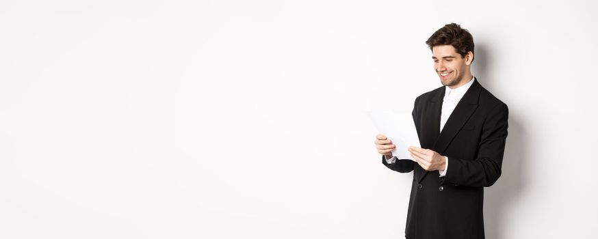 Image of attractive businessman in black suit, reading document and smiling, working on report, standing against white background.