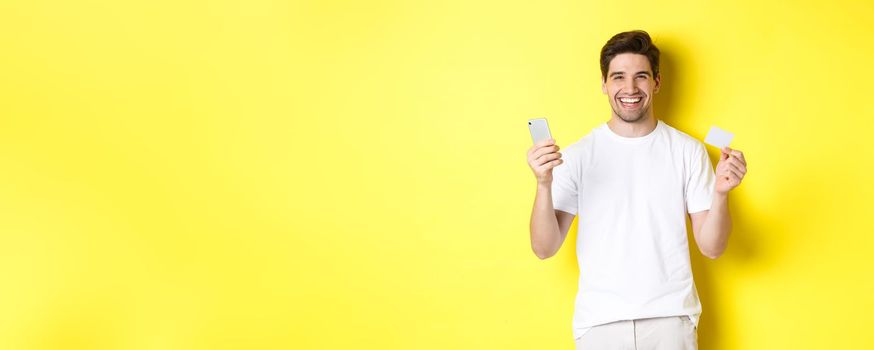 Happy young man shopping online in smartphone, holding credit card and smiling, standing over yellow background.