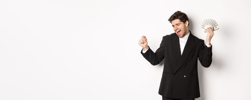 Portrait of handsome and successful businessman in suit, dancing with money, standing against white background.