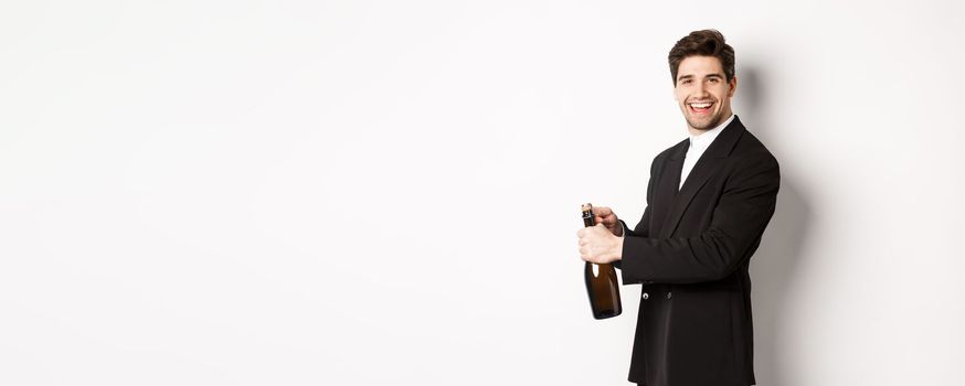 Handsome smiling man in trendy suit open a bottle of champagne, celebrating holidays, standing over white background.