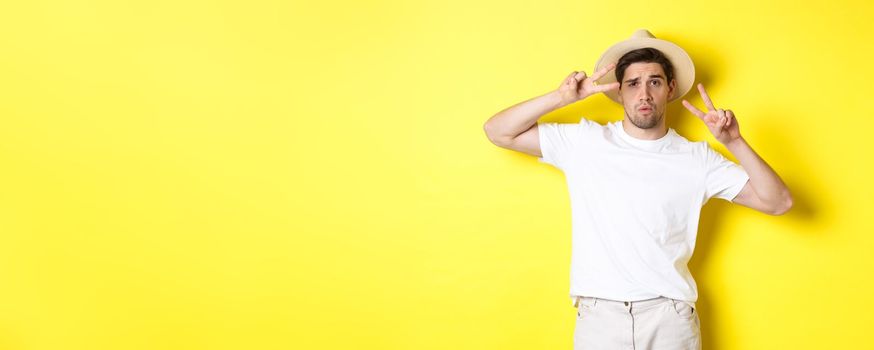 Concept of tourism and vacation. Cool guy taking photo on holidays, posing with peace signs and wearing straw hat, standing against yellow background.