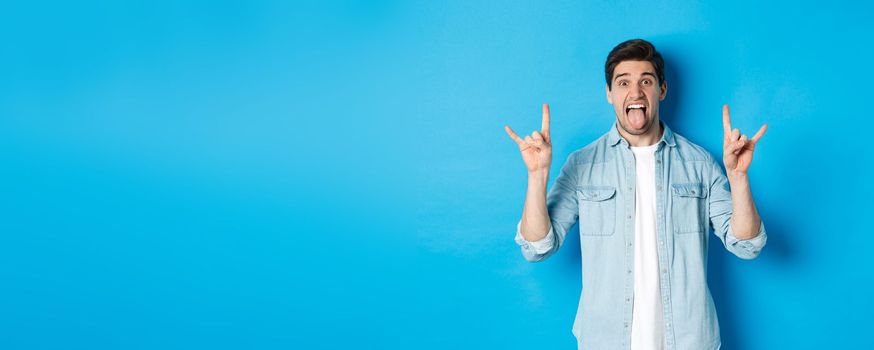 Cool guy having fun, enjoying concert, showing rock sign and tongue, standing over blue background.