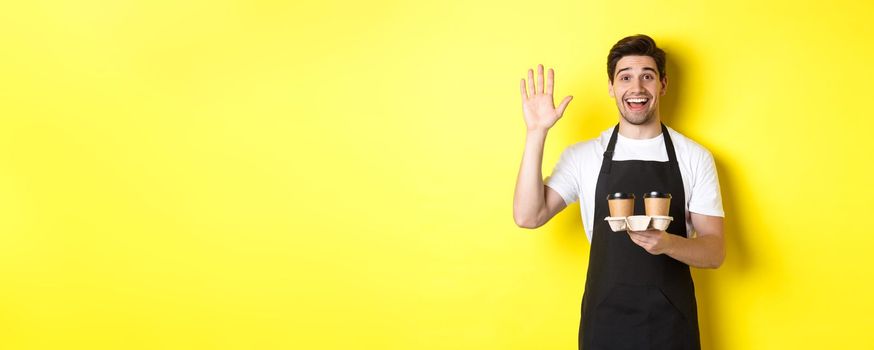 Friendly waiter in cafe waving hand at customer, holding takeaway coffee oder, standing against yellow background in black apron.