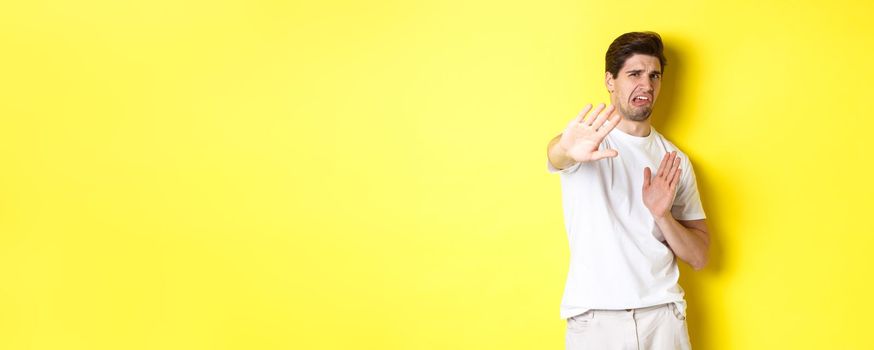 Disgusted man refusing, grimacing from dislike and aversion, begging to stop, standing in white t-shirt against yellow background.