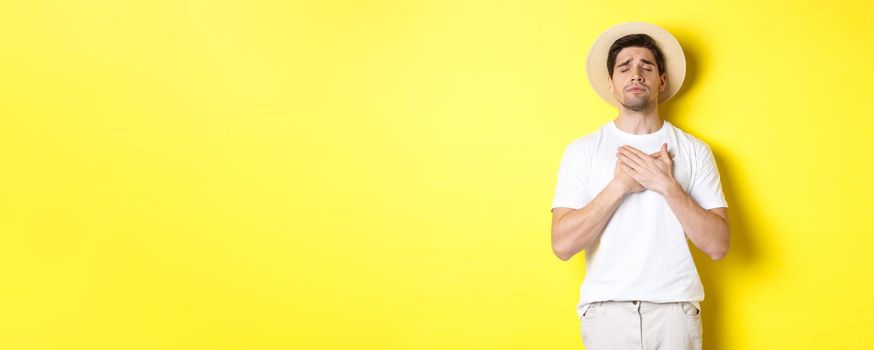 Concept of tourism and summer. Romantic man in straw hat looking nostalgic, close eyes and holding hands on heart, standing against yellow background.