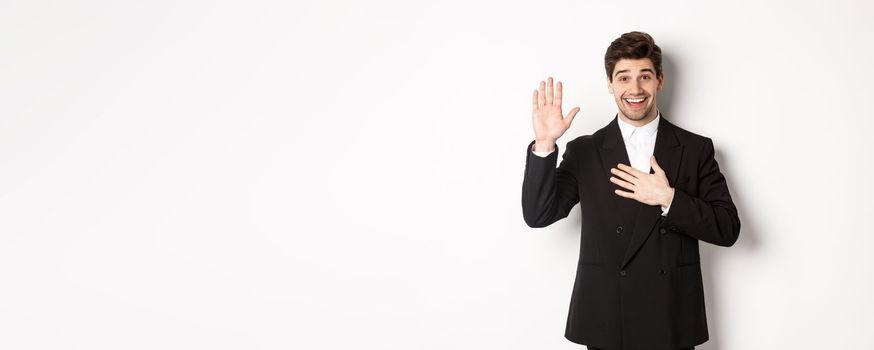 Image of handsome smiling guy telling the truth, raising on arm to promise something, standing over white background in black suit.