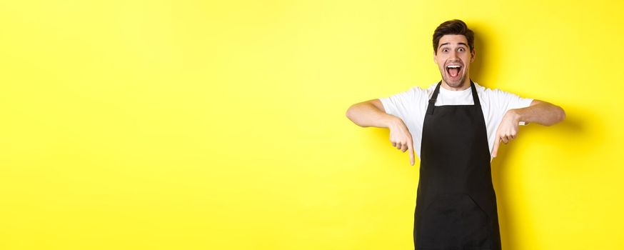Waiter, coffee shop barista in black apron pointing fingers down, looking amazed, standing over yellow background.