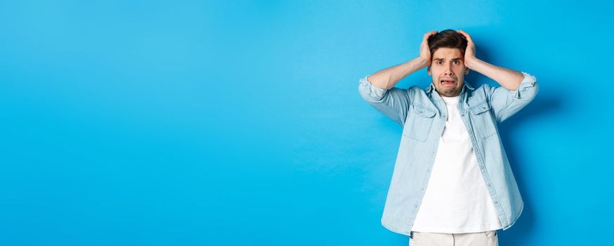 Image of man in panic holding hands on head, looking frustrated and anxious, standing against blue background.