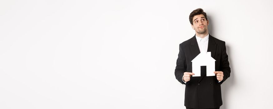 Image of handsome businessman in black suit, looking for home, holding house maket and gazing dreamy at upper right corner, standing against white background.