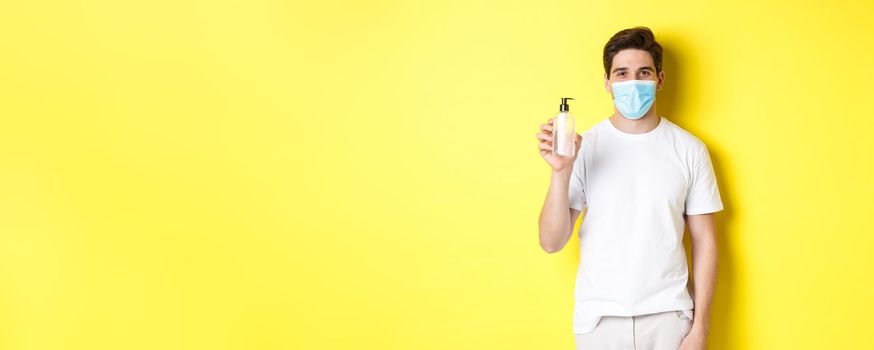 Concept of covid-19, quarantine and lifestyle. Young man in medical mask showing hand sanitizer, hands disinfection product, standing over yellow background.
