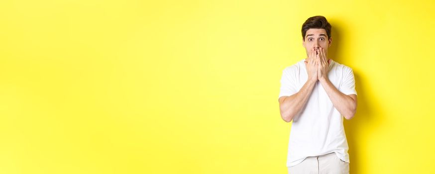 Handsome man looking shocked and speechless, holding hands on mouth, standing over yellow background.