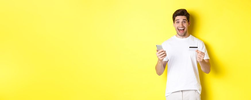 Happy male buyer holding smartphone and credit card, concept of online shopping in internet, standing over yellow background.