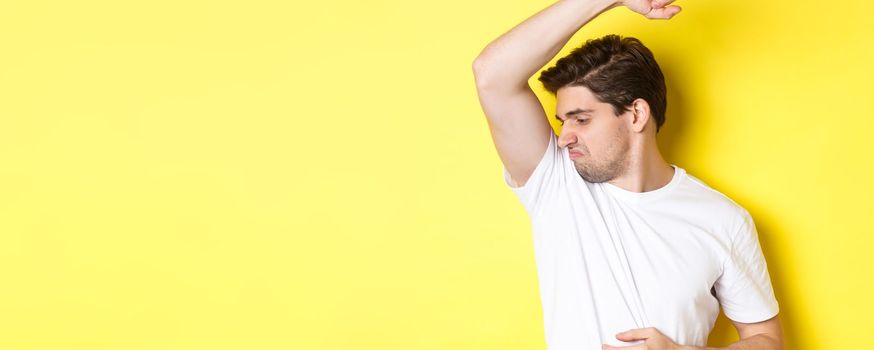 Man in sweat smelling his armpit, standing in white t-shirt and grimacing from stinky clothes.