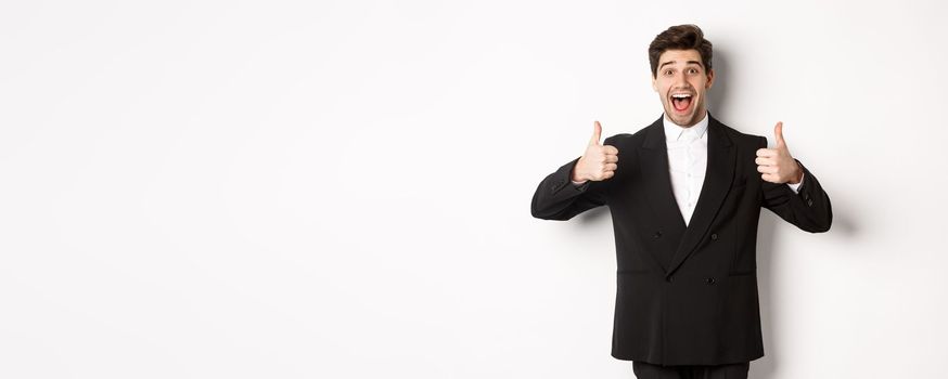 Concept of new year party, celebration and lifestyle. Portrait of amazed and pleased handsome man in black suit, showing thumbs-up, like product, approve something good, white background.