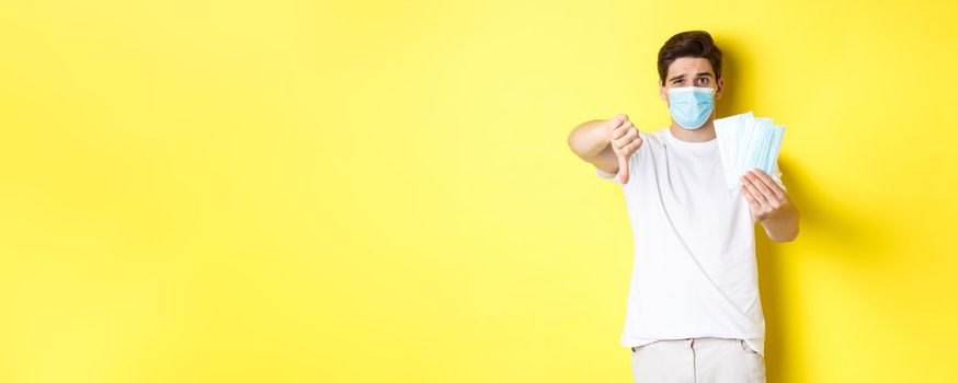 Concept of covid-19, quarantine and preventive measures. Man looking disappointed and showing thumb down, do not recommend bad medical masks, standing over yellow background.