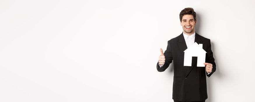 Portrait of confident real estate agent showing house maket and thumb-up, standing against white background.