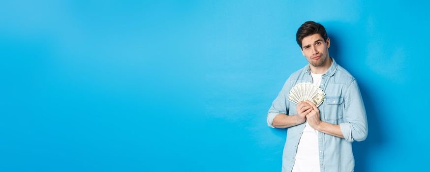 Handsome young man keeping money to himself, smiling and looking greedy, standing over blue background.