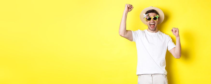 Concept of tourism and lifestyle. Happy man winning trip to resort, shouting yes and raising hands up, triumphing, wearing sunglasses and summer hat, yellow background.