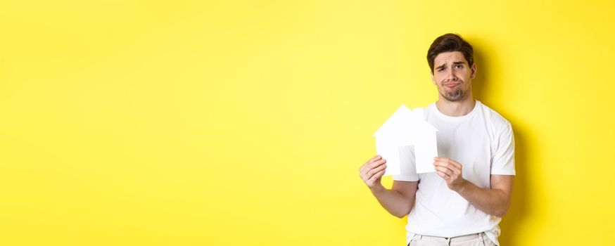 Real estate concept. Displeased young man showing paper house model and grimacing upset, standing over yellow background.