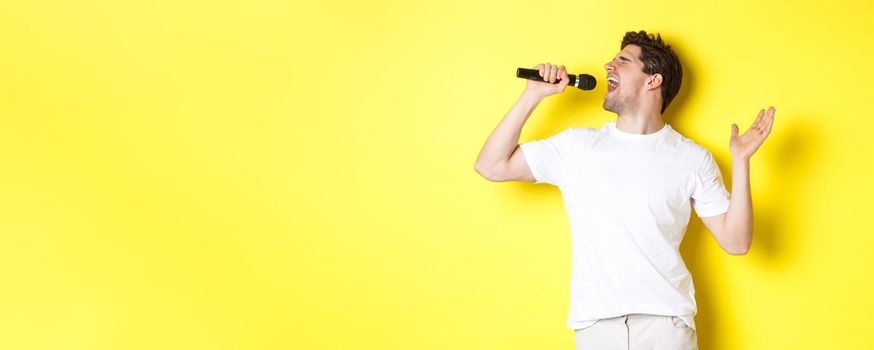 Young man singer holding microphone, reaching high note and singing karaoke, standing over yellow background.