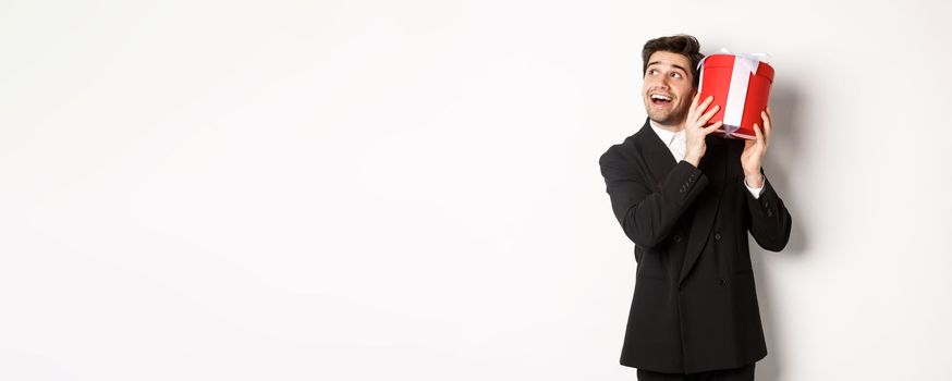 Concept of christmas holidays, celebration and lifestyle. Image of excited man enjoying new year, shaking gift box to guess what inside, standing against white background.