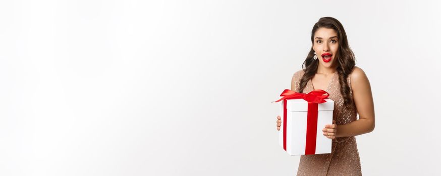 Merry Christmas. Image of beautiful woman in glamour dress receiving gift and looking surprised, celebrating New Year, standing over white background.