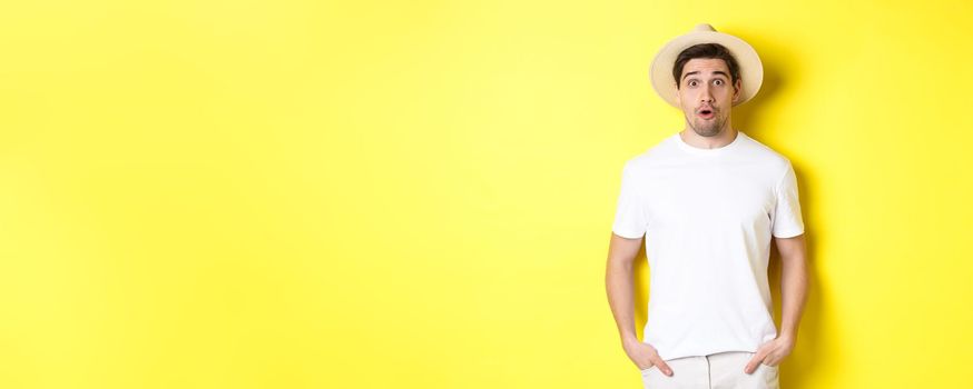 Concept of travelling and tourism. Surprised caucasian guy in straw hat, say wow, standing against yellow background.