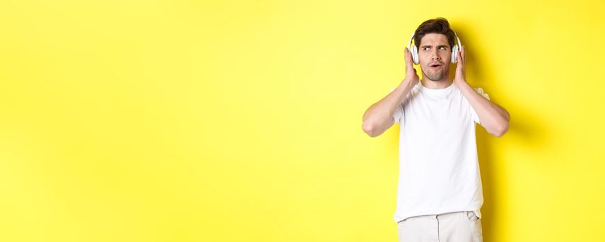 Intrigued guy enjoying tunes in headphones, listening closely to music in earphones, standing over yellow background.