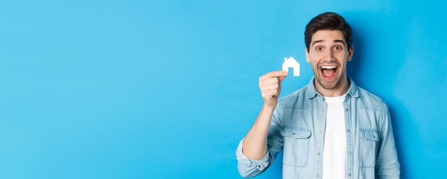 Real estate concept. Happy young man looking excited, found apartment, showing small house model, standing over blue background.