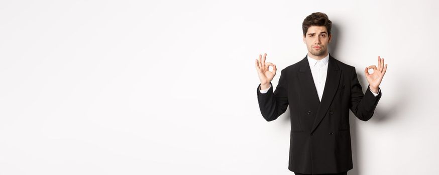 Concept of new year party, celebration and lifestyle. Portrait of confident good-looking man in black suit, showing okay sign and approve something, standing over white background.