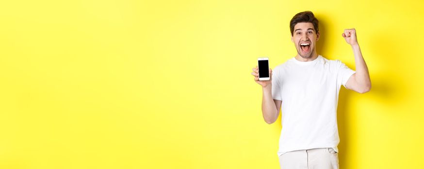 Cheerful guy showing smartphone screen, raising hand up and celebrating, triumphing over internet achievement, standing over yellow background.