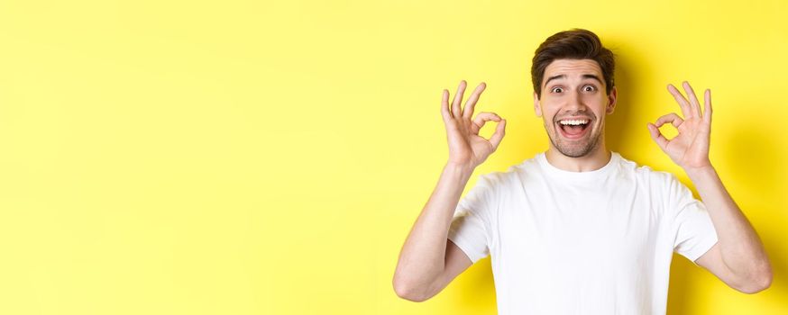 Amazed young man showing ok signs and smiling, recommending something good, standing over yellow background satisfied.