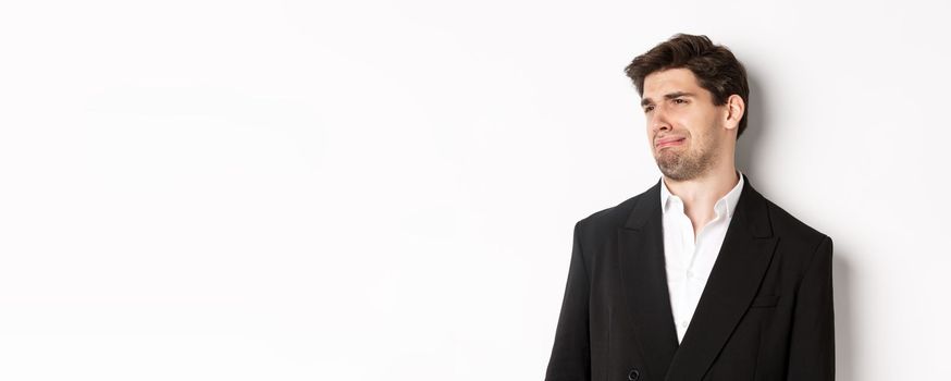 Close-up of disgusted young man in trendy suit, grimacing upset, looking left and standing against white background.