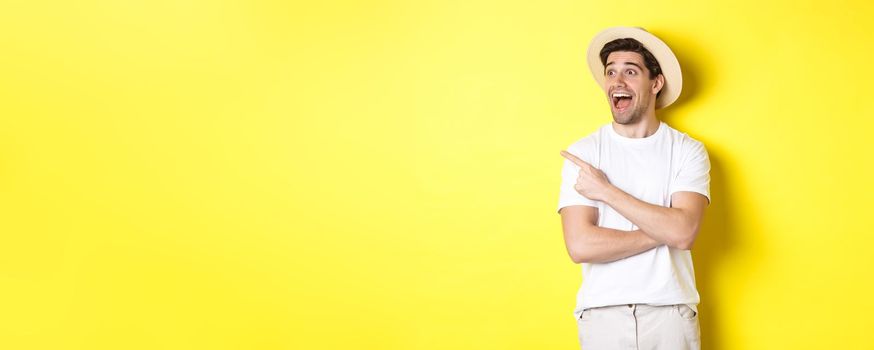 Concept of tourism and lifestyle. Excited handsome guy in straw hat checking out advertisement, pointing and looking at upper left corner logo, yellow background.