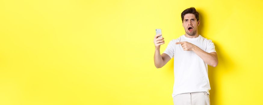 Confused man pointing finger at mobile phone screen, cant understand something, standing over yellow background. Copy space