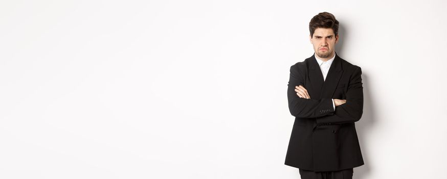 Portrait of angry handsome man in black suit, cross arms on chest and looking offended, frowning and pouting, being mad at someone, standing over white background.