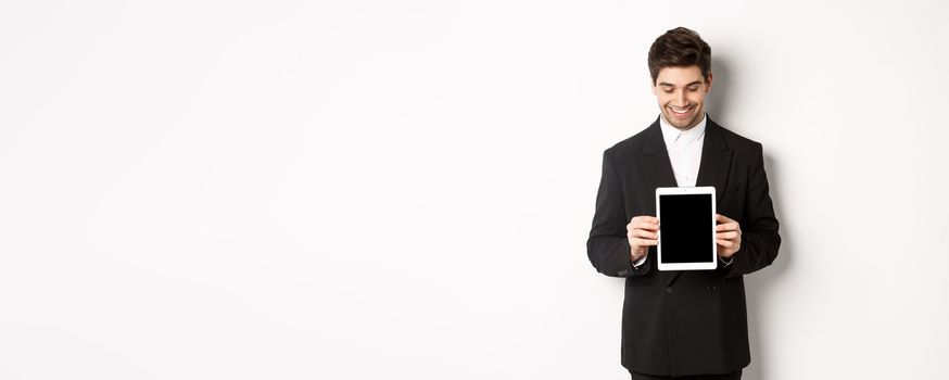 Image of good-looking male entrepreneur in black suit, looking down at digital tablet screen and showing advertisement, standing against white background.
