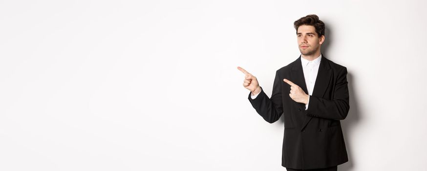 Portrait of serious handsome man in black suit, pointing and looking left at promo banner, showing advertisement on copy space, standing over white background.