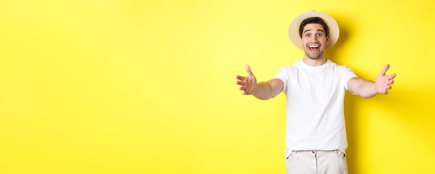 Concept of tourism and summer. Happy and friendly man traveller reaching hands for hug, greeting or welcome you, standing over yellow background.