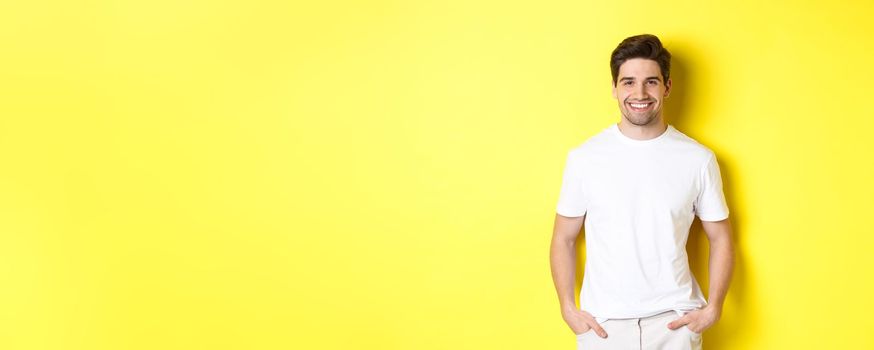 Young handsome man smiling at camera, holding hands in pockets, standing against yellow background.
