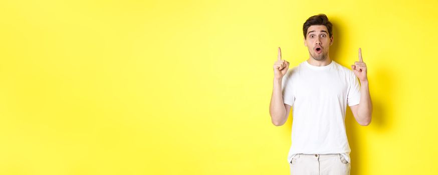 Surprised handsome guy in white t-shirt, pointing fingers up, interested about advertisement, standing against yellow background.