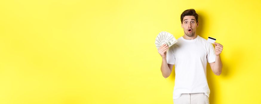 Amazed man holding credit card and money, looking surprised, standing over yellow background.