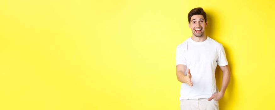 Friendly man greeting you with handshake, smiling amused, saying hello, standing over yellow background.