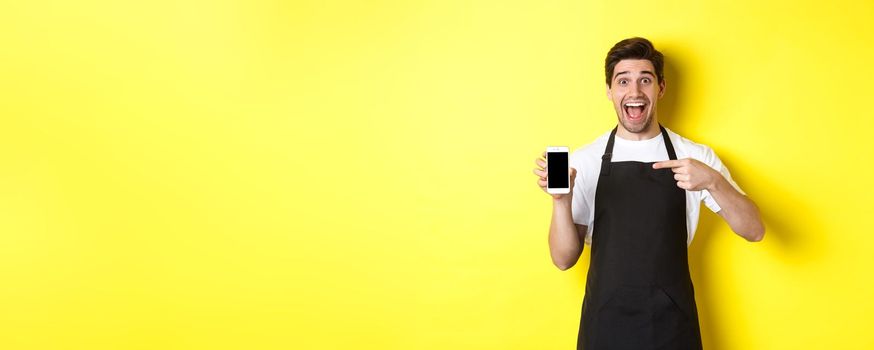 Handsome barista in black apron pointing finger at mobile screen, showing app and smiling, standing over yellow background.