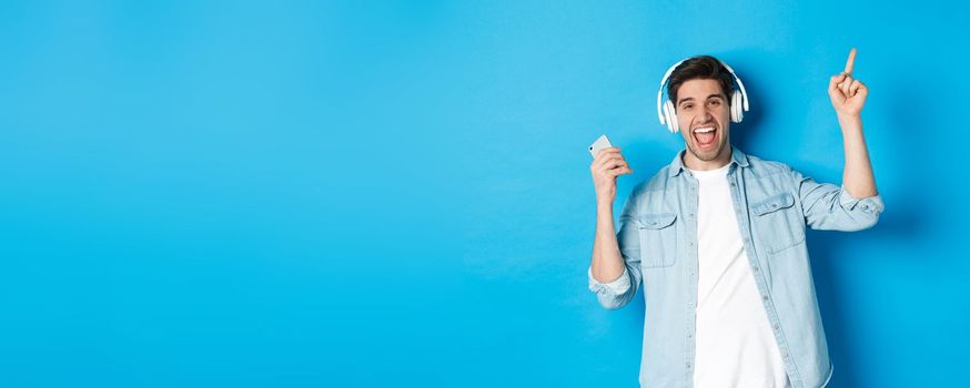 Joyful handsome man dancing with smartphone, listening music in headphones and pointing finger up, standing over blue background.